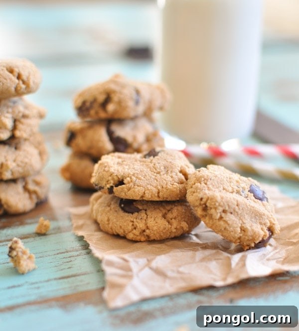 Small Paleo Chocolate Chip Cookies stacked on top of each other placed on brown parchment paper with a glass of milk in the background.