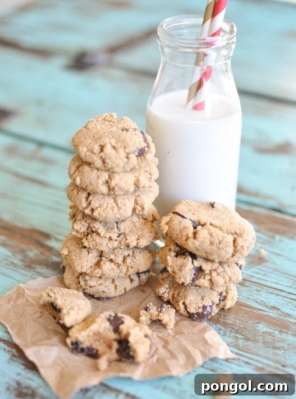 Paleo Chocolate Chip Cookies stacked on top of each other on brown parchment paper leaning against a glass of milk with two straws sticking out.