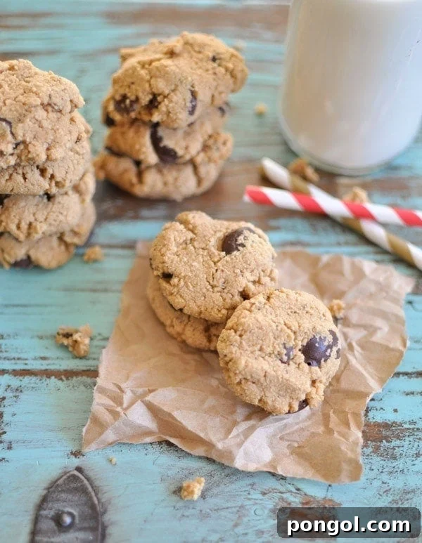 Paleo Chocolate Chip Cookies stacked on top of each other on brown parchment paper with some straws and a glass of milk in the background.