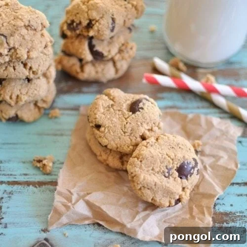 chocolate chip almond cookies stacked up on a piece of brown parchment paper with a glass of milk in the background.