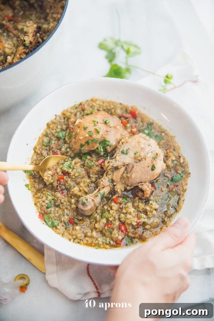A bowl of healthy Spanish chicken and cauliflower rice stew being held, with a pot of the stew in the background. The stew is garnished with fresh cilantro and a few olives, emphasizing its vibrant appeal.
