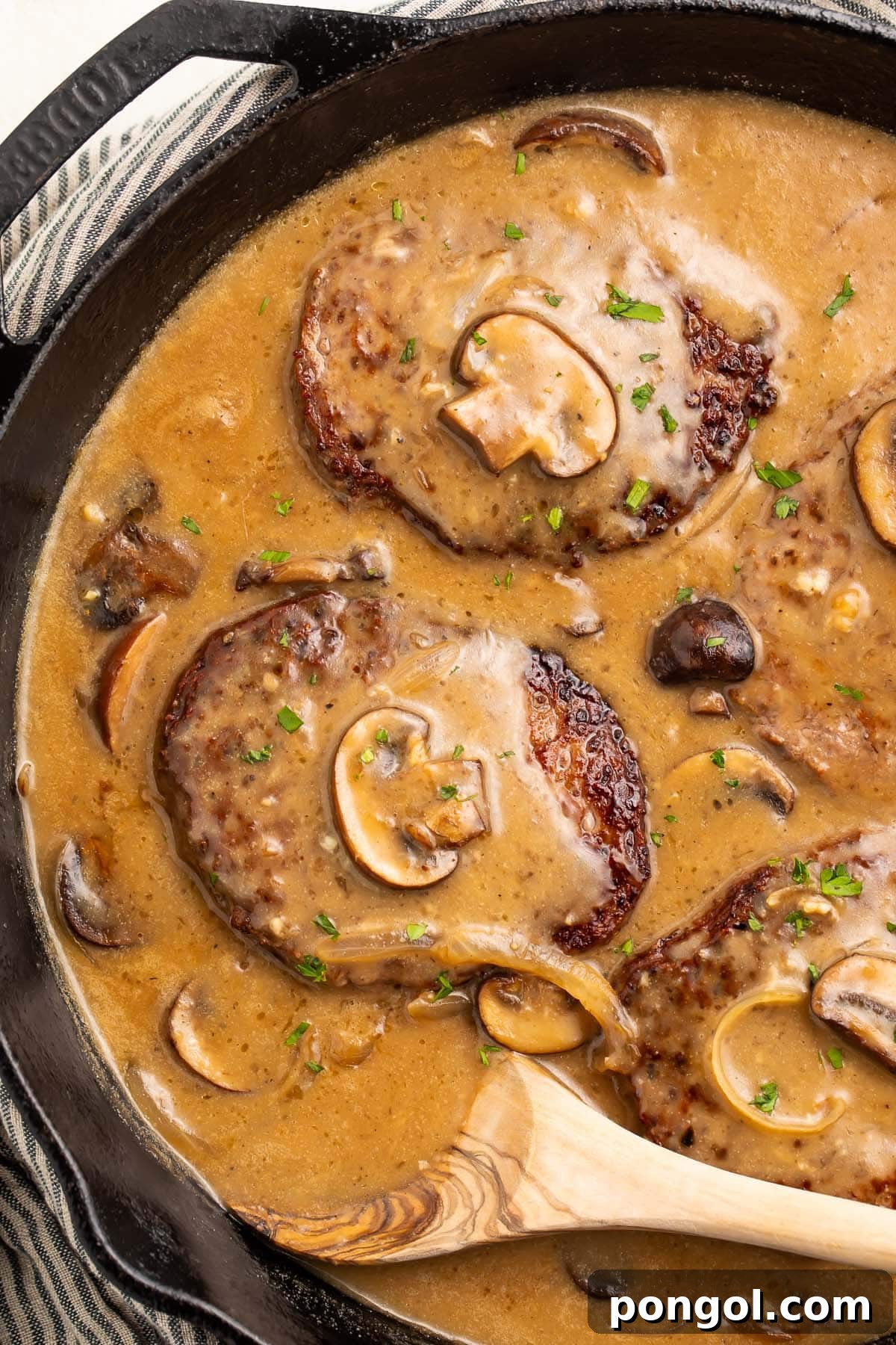 Four Whole30 salisbury steaks in a cast-iron skillet with plenty of salisbury sauce, mushrooms, and a wooden spoon.