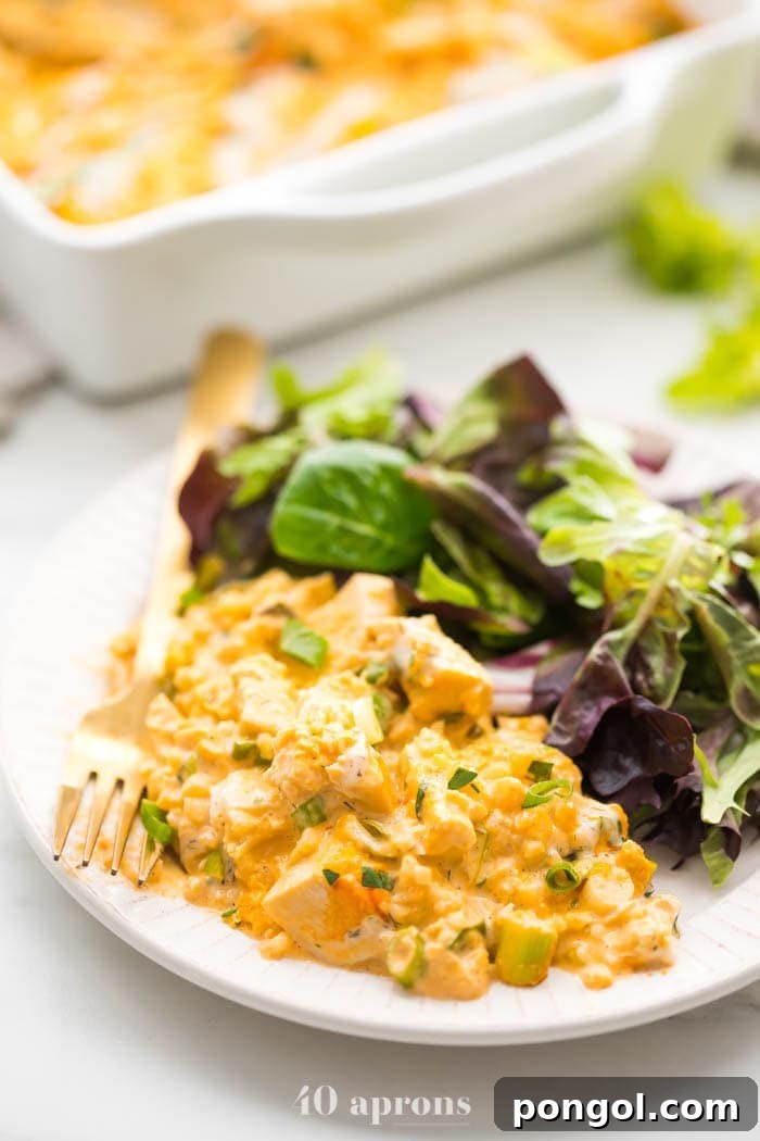 A serving of Paleo Buffalo Chicken Casserole on a white plate, accompanied by a fresh green salad, with the full baking dish visible in the soft-focused background, emphasizing a wholesome meal.