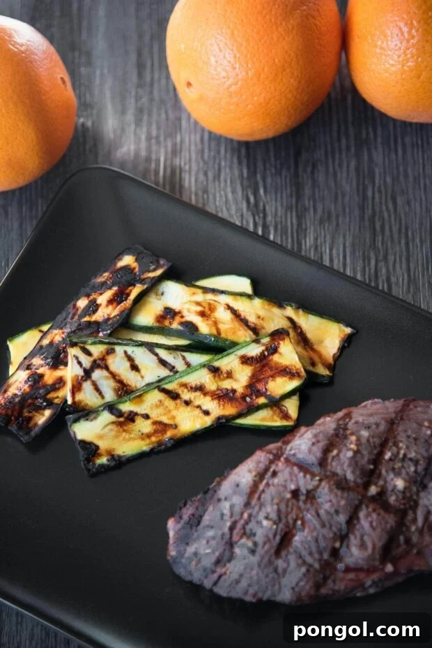 Sweet & Smoky Orange Glazed Zucchini (Paleo, Gluten Free) 2 Overhead shot of grill-marked orange glazed grilled zucchini slices arranged on a black plate next to a steak with some oranges in the background.