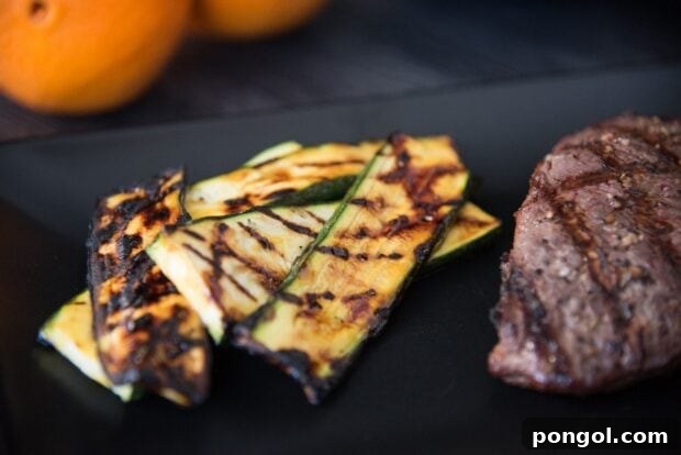 Sweet & Smoky Orange Glazed Zucchini (Paleo, Gluten Free) 8 Close-up of grill-marked orange glazed zucchini slices arranged on a black plate next to a steak with oranges in the background.