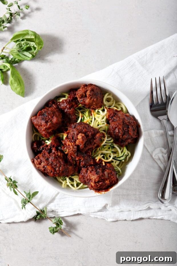 Whole30 Spaghetti and Meatballs served over zucchini noodles in a white bowl on a white tablecloth with some fresh herbs in the background.