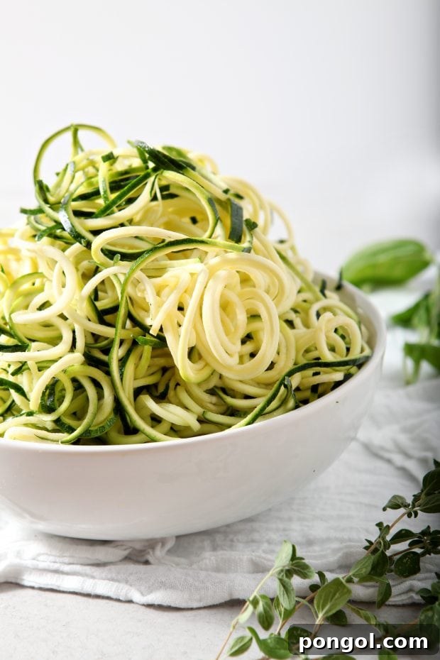 side view of raw zucchini noodles in a white bowl on a white tablecloth with some fresh herbs in the background
