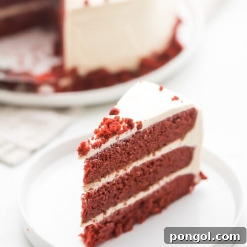 Slice of gluten free red velvet layer cake with paleo cream cheese frosting on a white plate with whole cake in background
