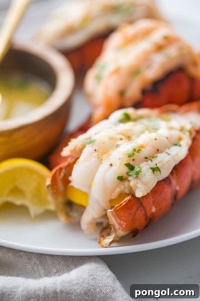 Broiled lobster tails butterflied on a plate with parsley and a lemon wedge next to a wooden bowl of garlic butter dipping sauce