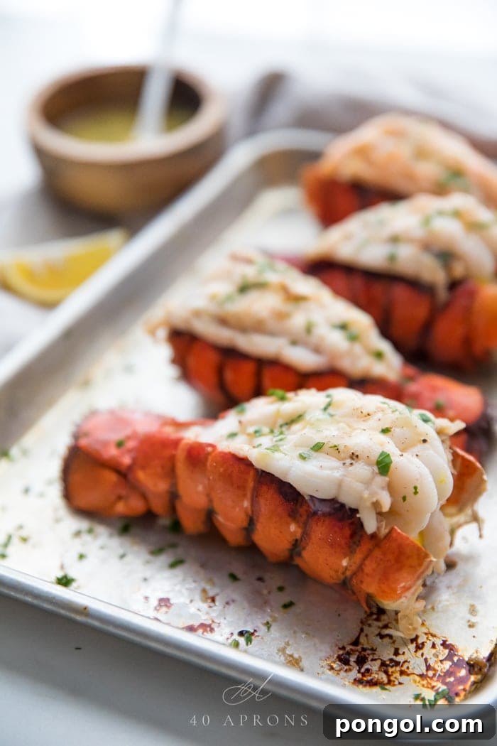 Broiled butterflied lobster tails on a baking sheet with a lemon wedge and small wooden bowl of garlic butter dipping sauce in the background