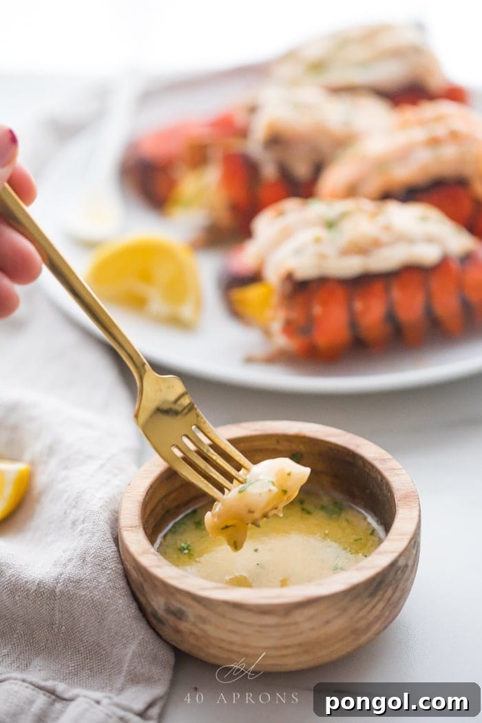 A piece of broiled lobster tail on a fork being dipped into a small wooden bowl of garlic butter dipping sauce with a plate of broiled lobster tails on a plate in the background