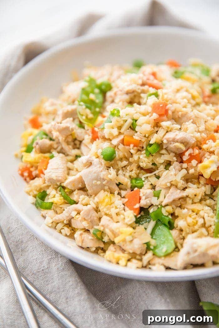A white bowl of cauliflower fried rice with chicken, peas, carrots with a grey linen and silver chopsticks to the side