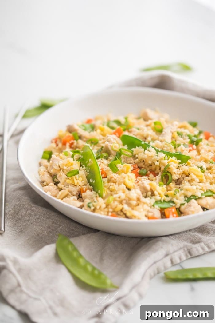A white bowl of cauliflower fried rice with chicken, peas, carrots with a grey linen and silver chopsticks to the side
