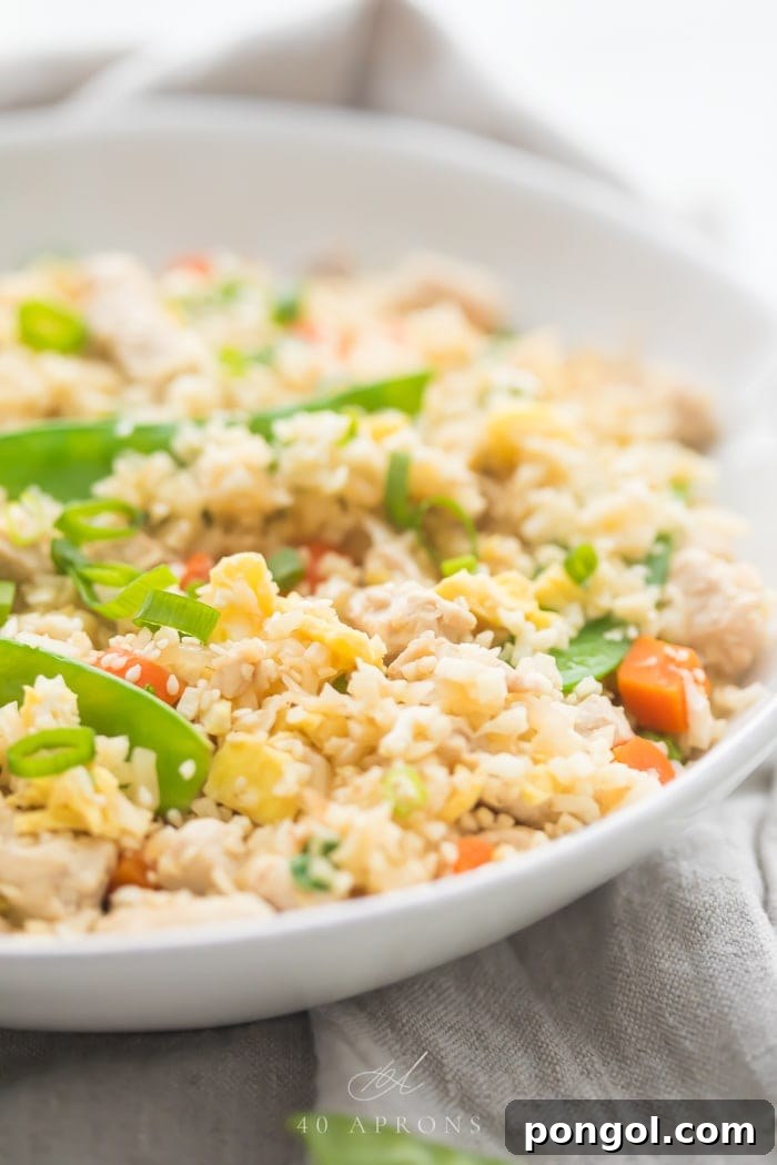 A white bowl of cauliflower fried rice with chicken, peas, carrots with a grey linen and silver chopsticks to the side