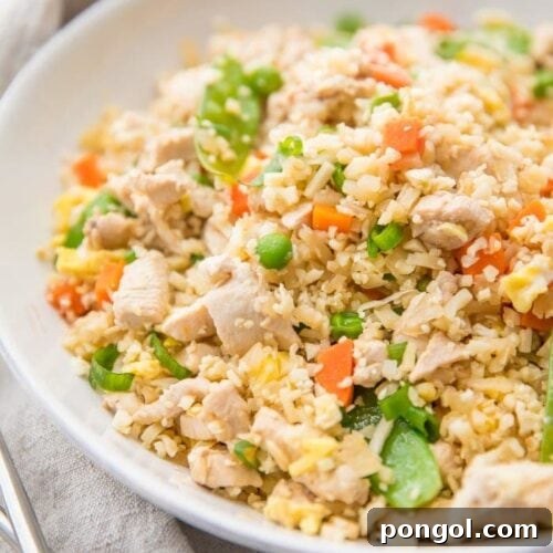 A white bowl of cauliflower fried rice with chicken, peas, carrots with a grey linen and silver chopsticks to the side