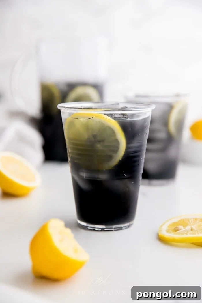 A glass of black charcoal lemonade with lemon slice, with glass and pitcher in background, highlighting its unique dark color