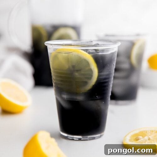 A glass of black charcoal lemonade with lemon slice, with glass and pitcher in background