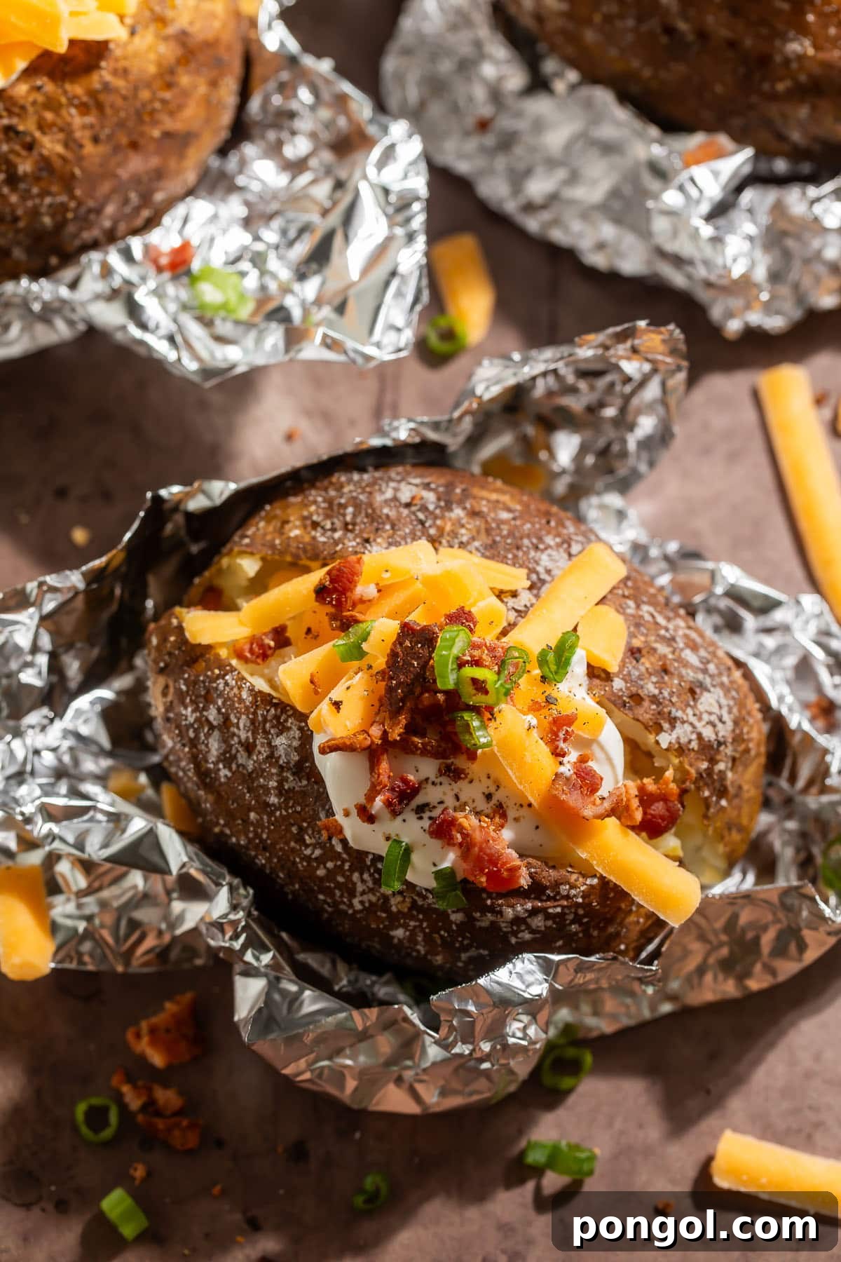 Foil-wrapped baked potatoes, cooked on a grill, split open and topped with shredded cheddar, a dollop of sour cream, and chopped green onions, showcasing a delicious salt crust and fluffy interior.