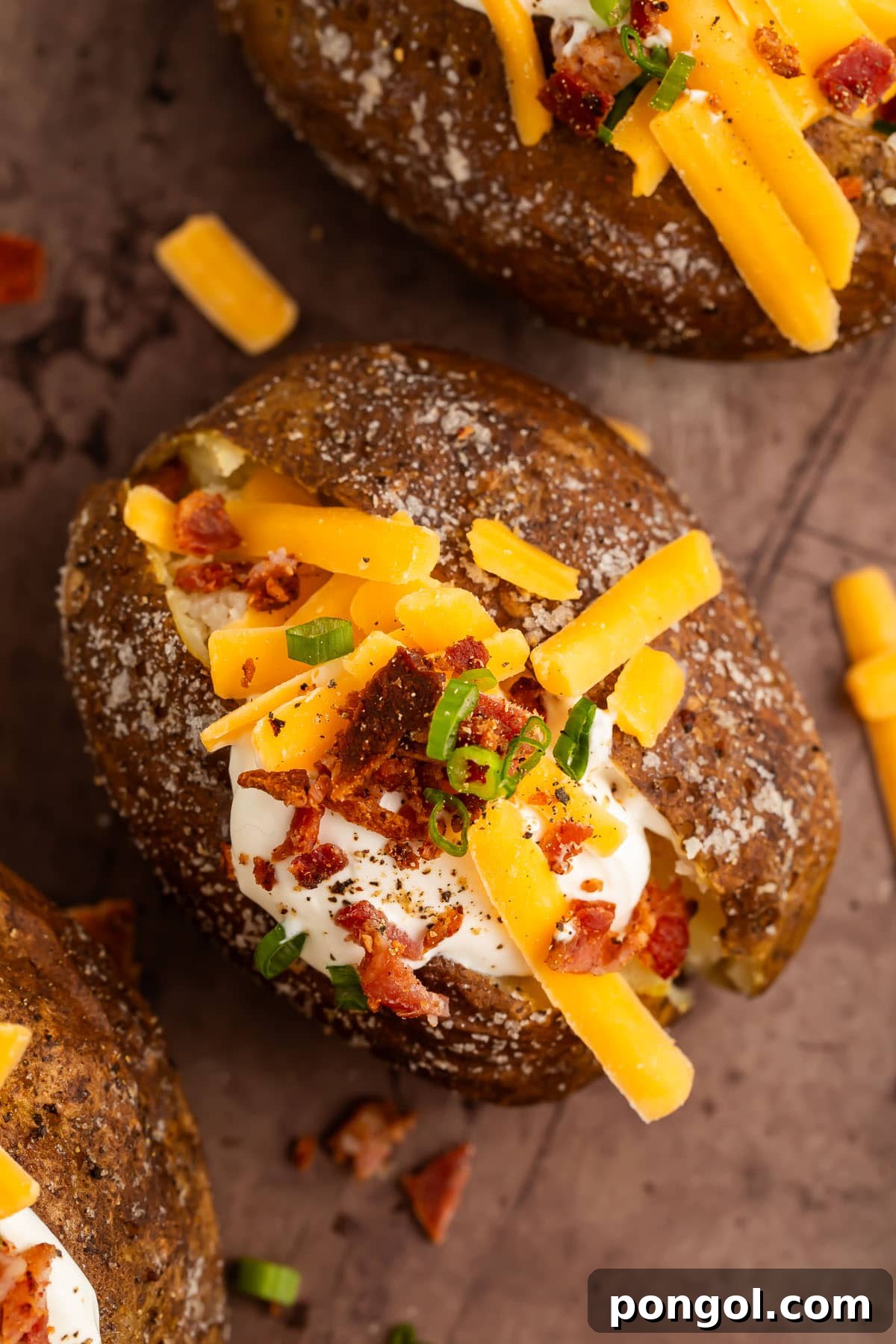 Two grilled baked potatoes, split open and generously topped with sour cream, cheddar cheese, chopped green onions, and crispy bacon bits on a wooden board.