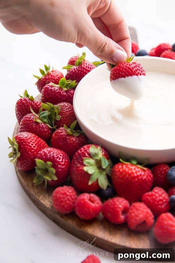 A bowl of creamy vanilla yogurt fruit dip with a fresh strawberry being dipped into it, surrounded by vibrant berries.