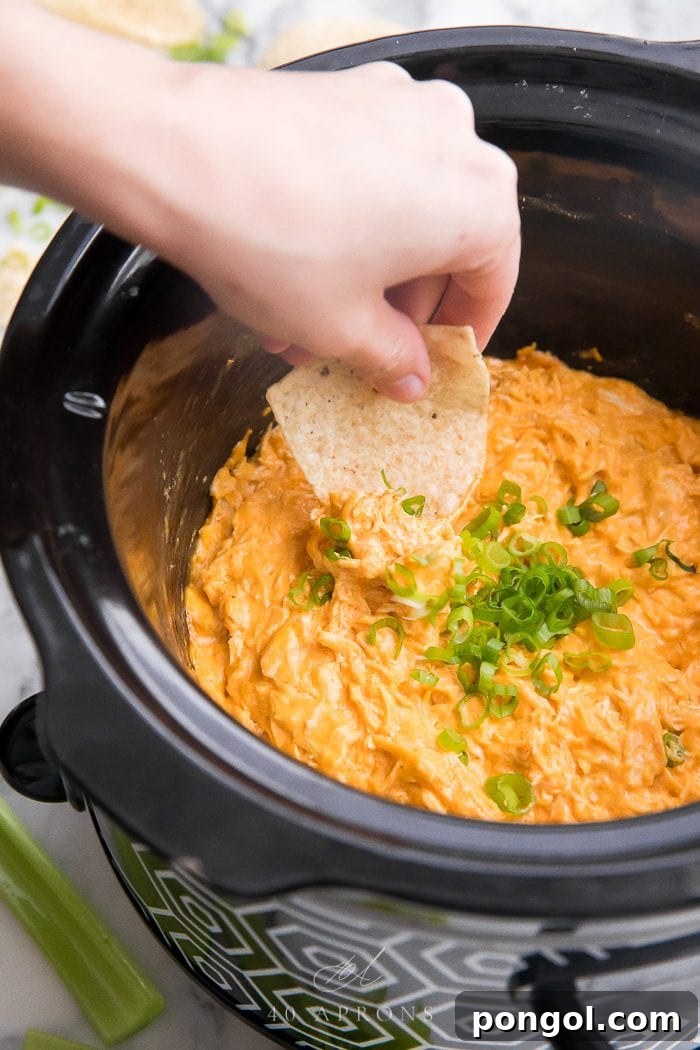 A tortilla chip scooping up a generous portion of warm, bubbly buffalo chicken dip from a slow cooker.