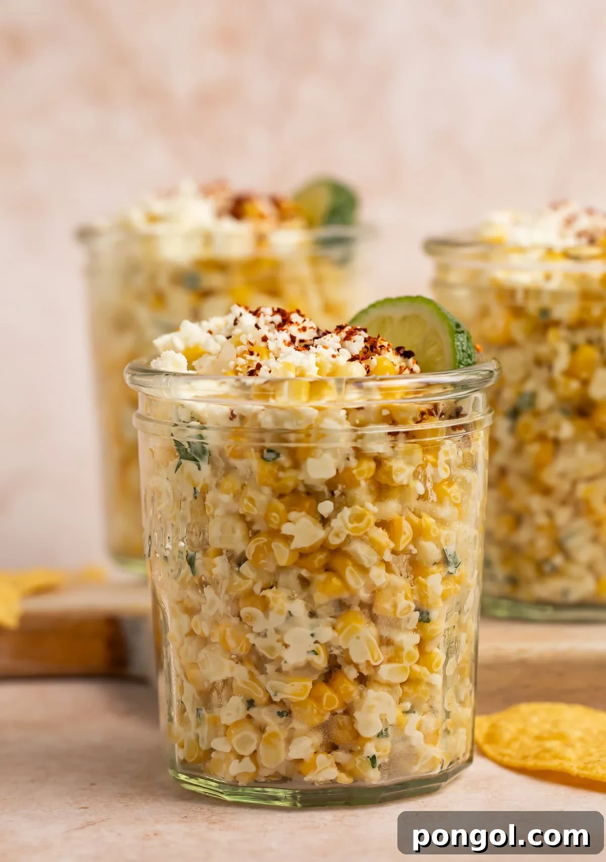 Several glass jars of vibrant Mexican street corn salad (esquites) arranged on a blue striped napkin with tortilla chips beside them, ready for serving.