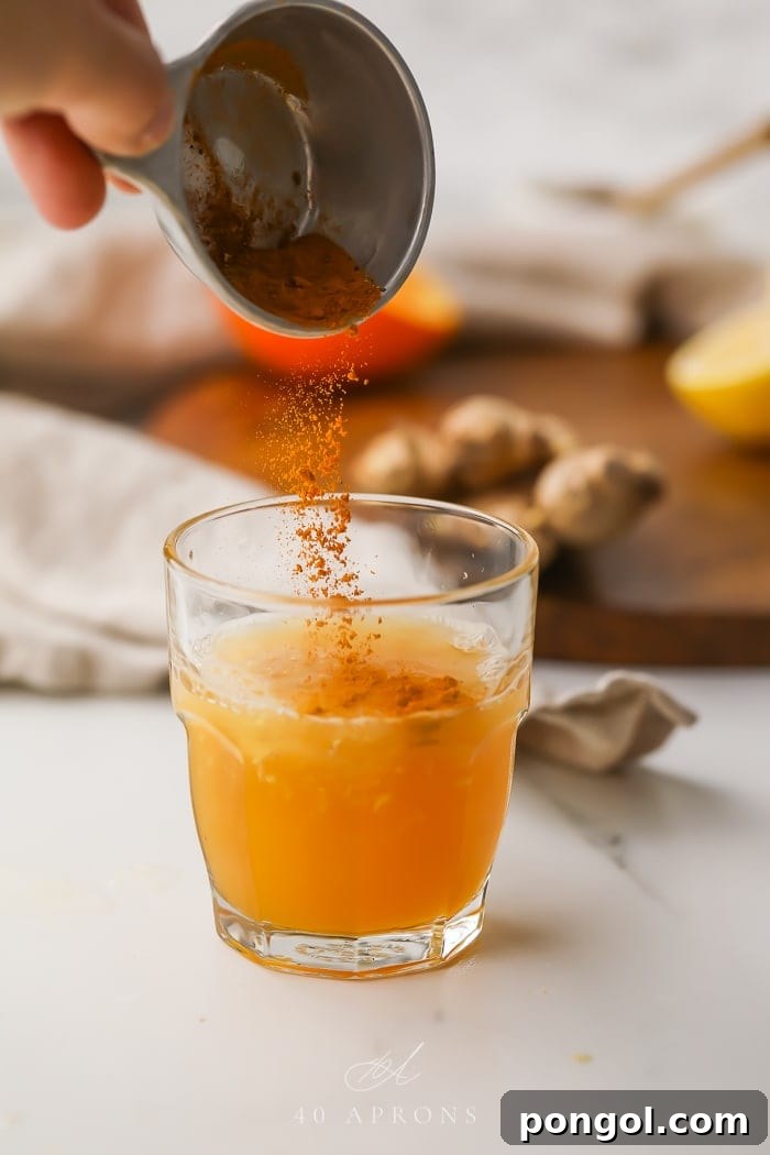 Various spices, including turmeric and cayenne pepper, being carefully poured from small bowls into a glass containing the liquid base of the immune booster shot.