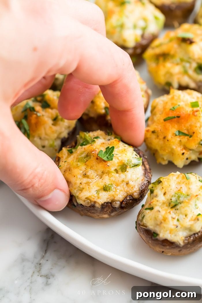 A hand picking up one of the golden-brown crab stuffed mushrooms from a white plate