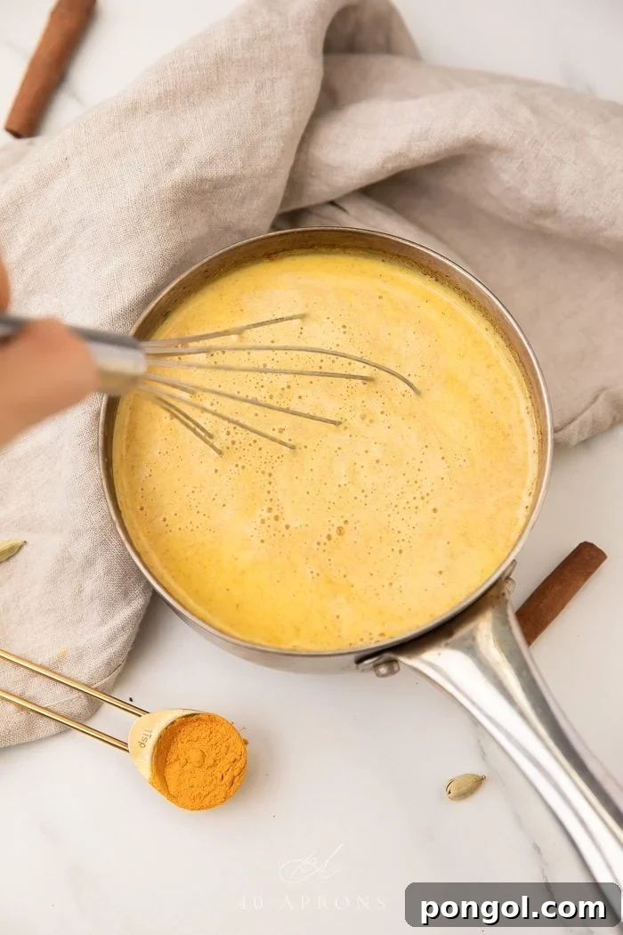 A whisk stirring creamy golden milk in a stainless steel saucepan on a stovetop, demonstrating the gentle heating process for optimal flavor infusion.