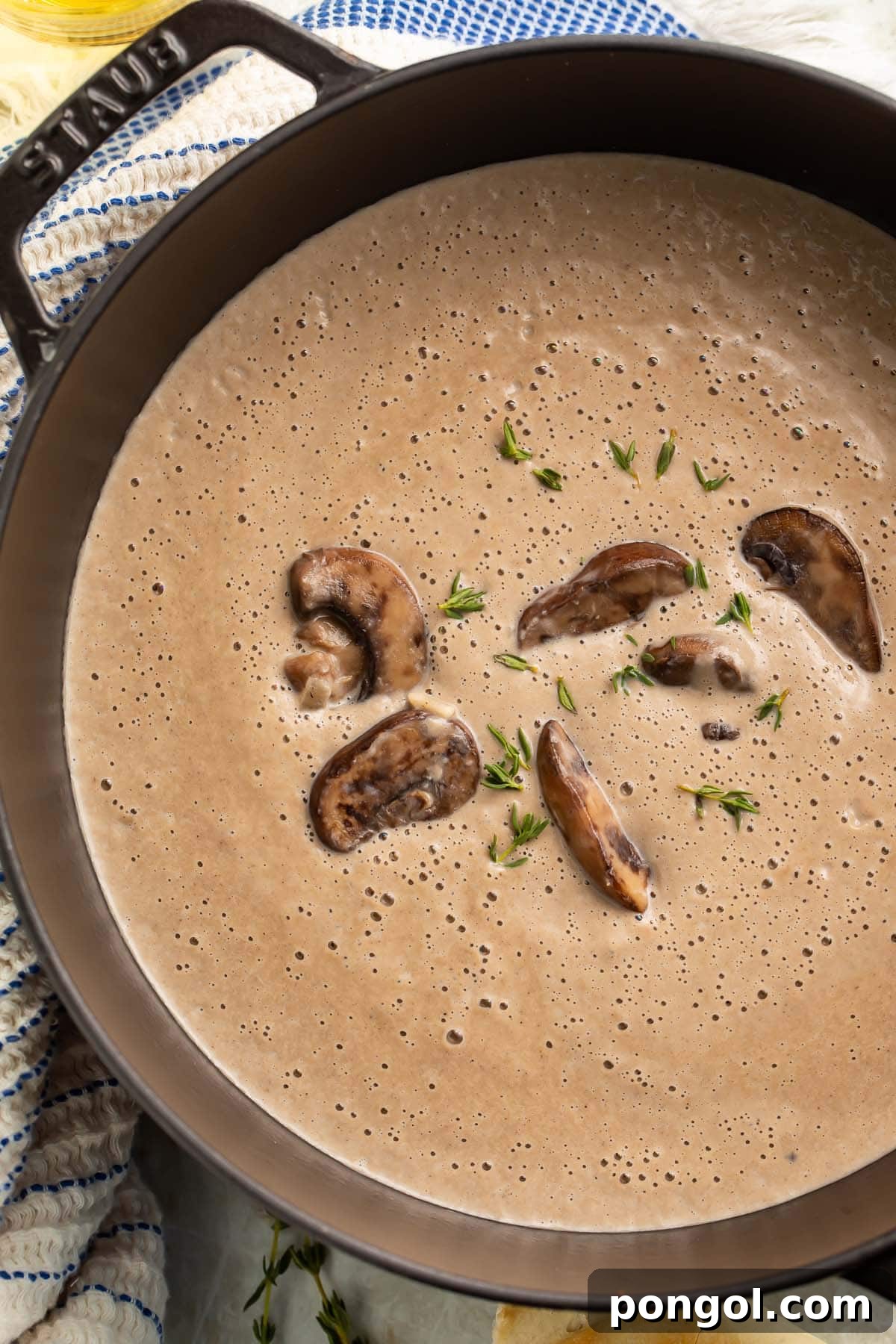 Overhead view of rich, thick, grayish-brown homemade cream of mushroom soup in a large round black pot, garnished with fresh herbs.