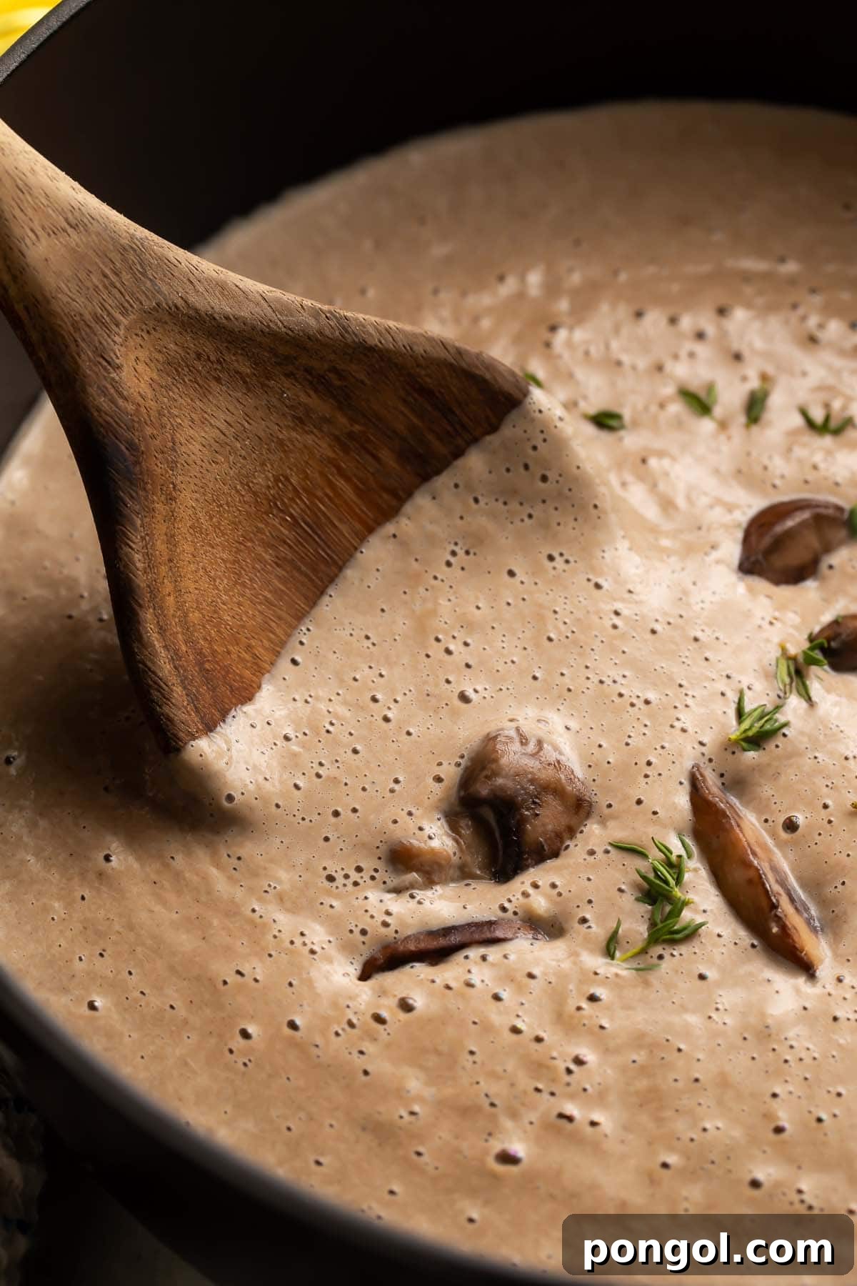 A close-up photo of a large wooden spoon stirring a creamy, thick cream of mushroom soup in a large black pot, steam rising appetizingly.