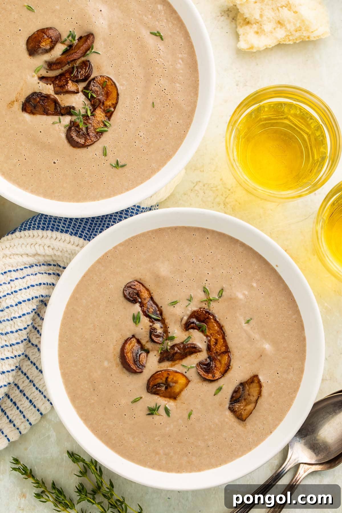 Overhead shot of two white bowls of grayish-brown cream of mushroom soup, perfectly garnished, on a rustic wooden table with a napkin and a glass of white wine.