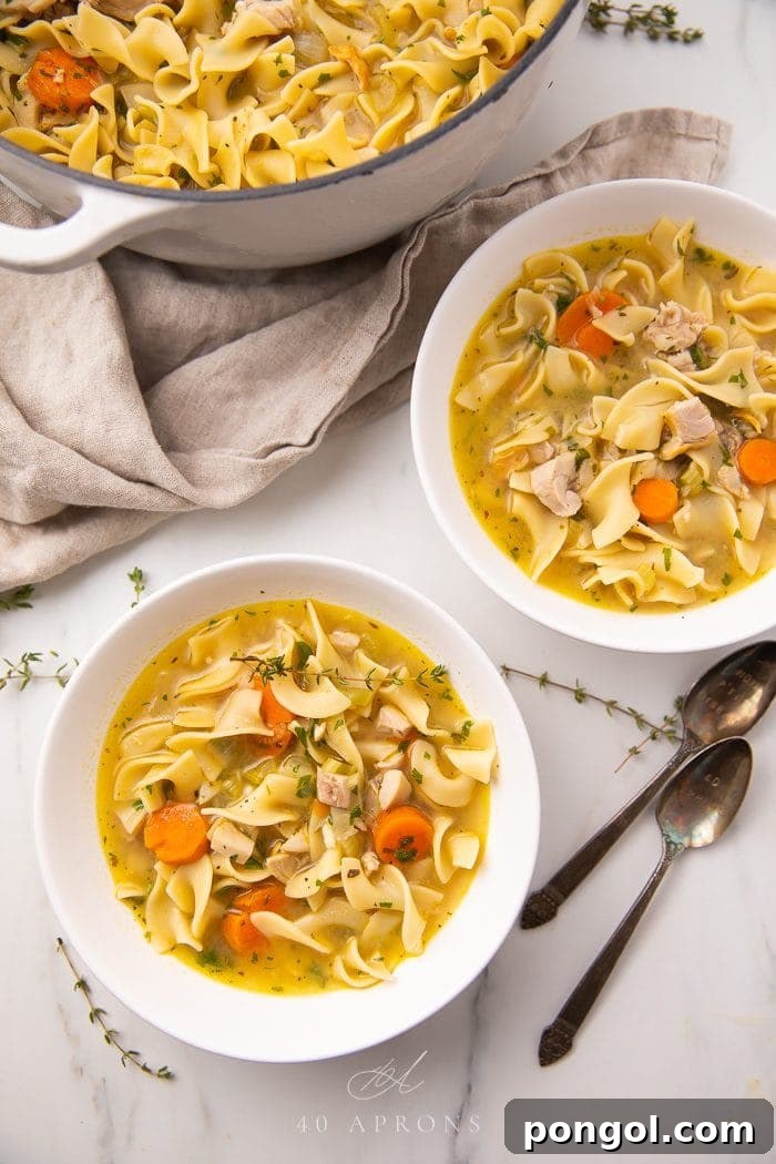Two white bowls and a Dutch oven filled with hearty leftover turkey noodle soup, garnished with fresh thyme on a elegant marble surface. The soup features tender turkey, vibrant carrots, and classic egg noodles.