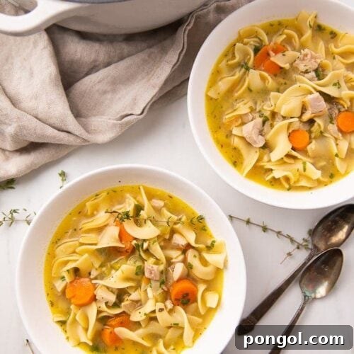 Two white bowls and a Dutch oven filled with leftover turkey noodle soup with carrots and egg noodles, served with fresh thyme on a marble surface.