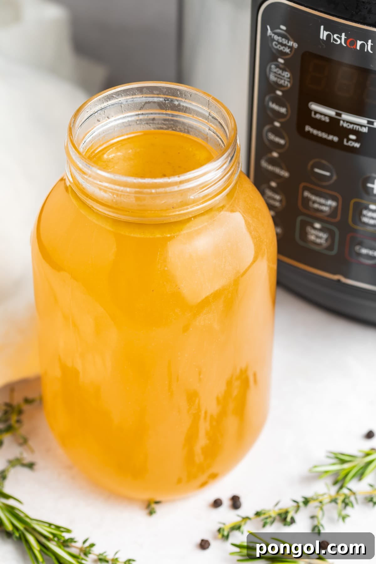 A clear mason jar filled with golden yellow Instant Pot turkey stock, set against a backdrop of an Instant Pot pressure cooker and fresh aromatic herbs, highlighting its rich color and natural ingredients.