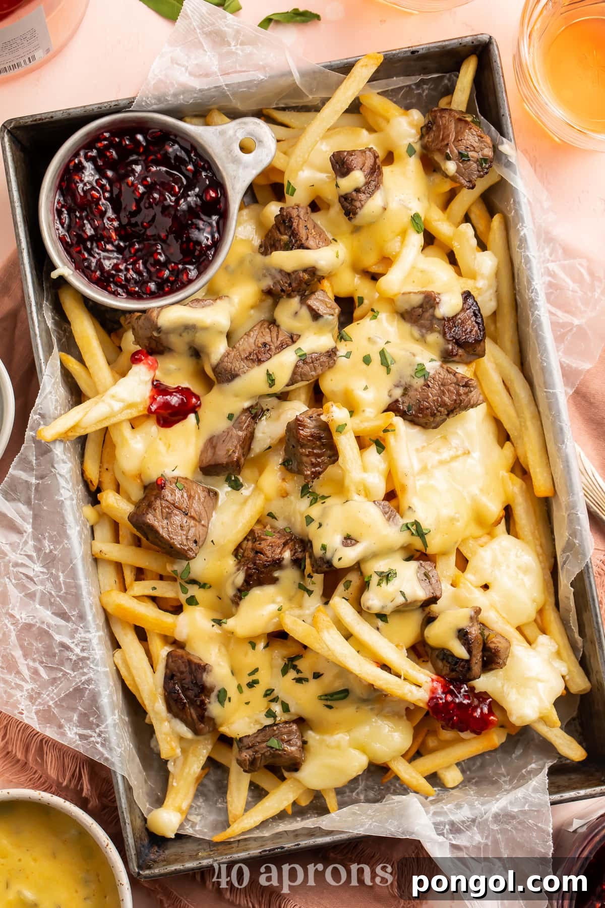 An overhead, angled shot of an indulgent dish of Baked Brie and Steak Cheese Fries, presented in a rectangular deep pan, served with a vibrant side of raspberry preserves.