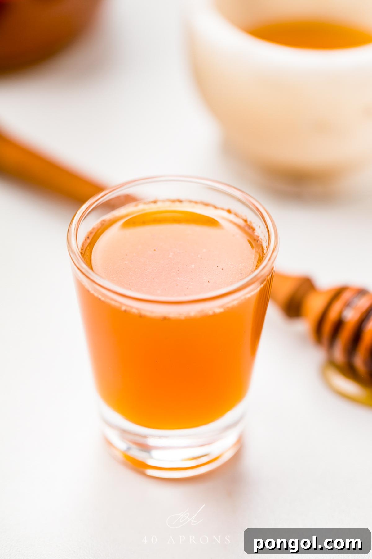 A clear glass shotglass holding an orange liquid peppered with cinnamon, in front of a honey wand dripping with honey, symbolizing the natural ingredients of an Apple Cider Vinegar Honey Shot.