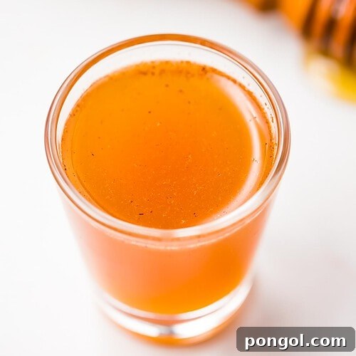 A clear glass shotglass holding an orange liquid peppered with cinnamon, in front of a honey wand dripping with honey.