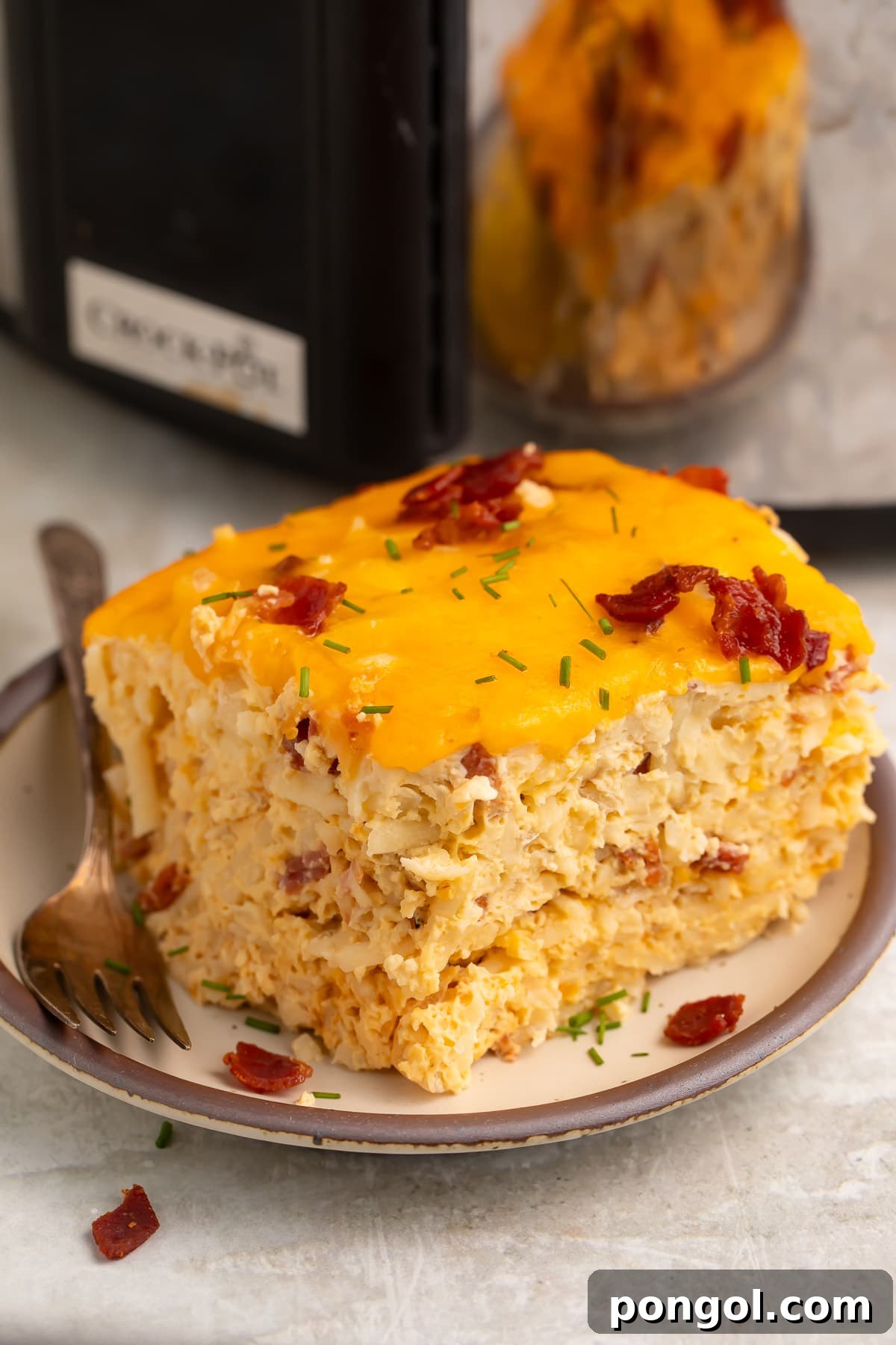 A square, multi-layered slice of Crockpot breakfast casserole on a plate in front of a slow cooker. The casserole is golden brown with visible layers of egg, potato, and cheese.