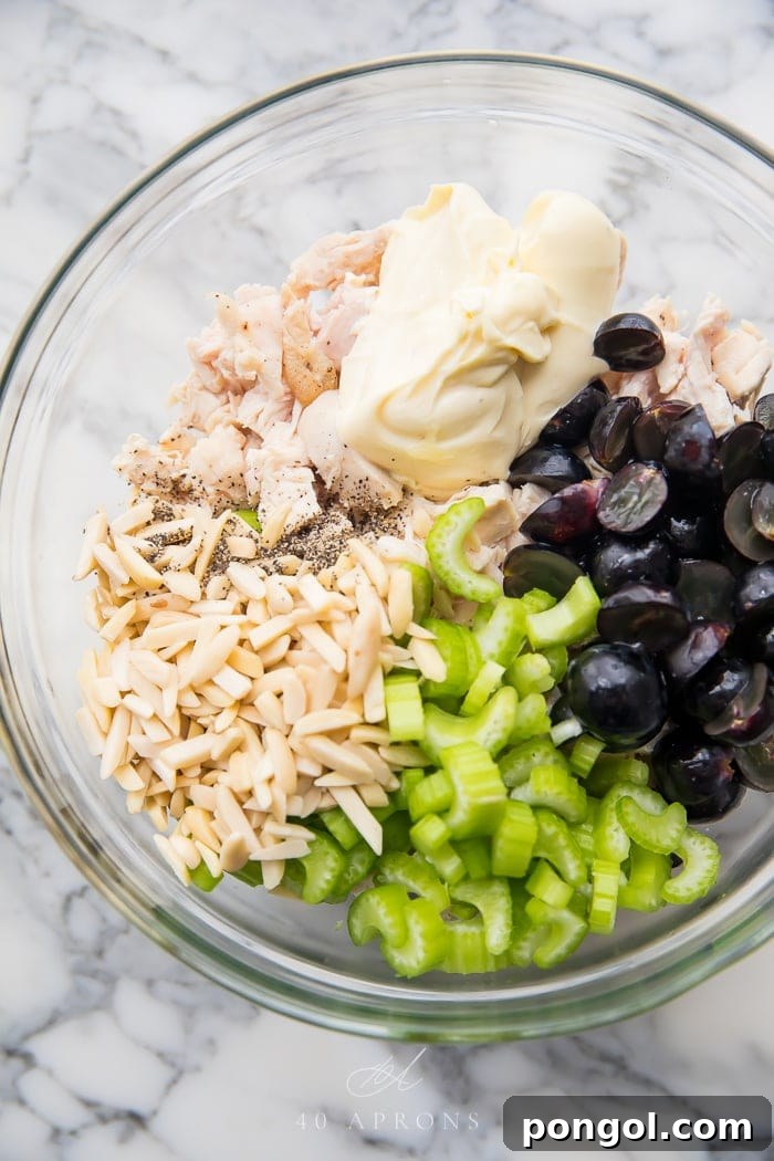 Fresh ingredients for a Whole30 chicken salad, including cooked chicken, grapes, celery, and almonds, displayed in a clear glass bowl, ready for mixing.