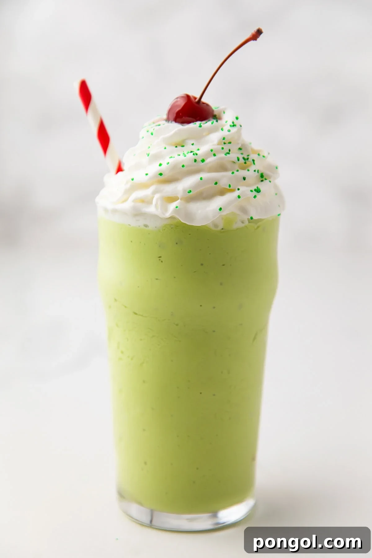 Emerald Vegan Mint Shake 3 A close-up of a green dairy-free shamrock shake copycat, topped generously with dairy-free whipped cream, a bright red cherry, and a festive red-and-white striped straw.