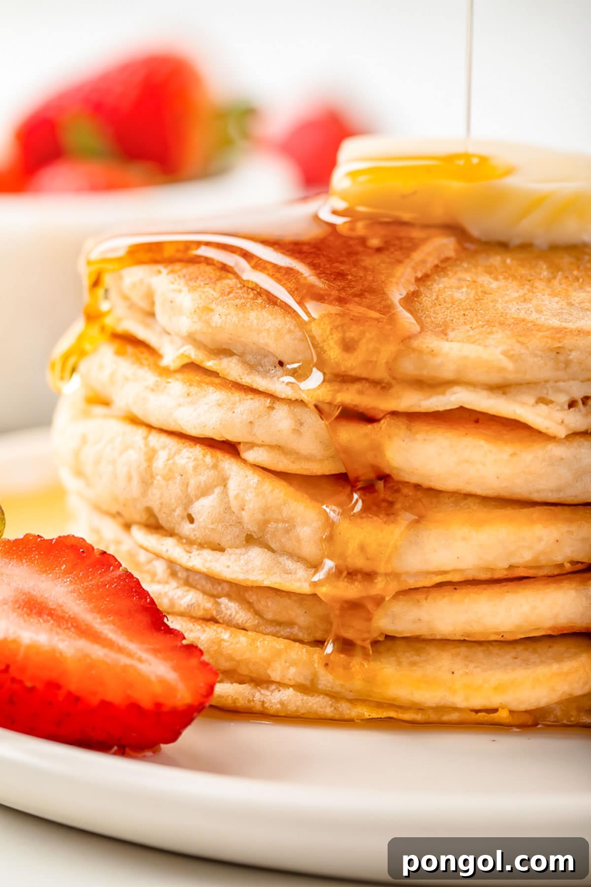 Syrup being poured over a stack of fluffy gluten-free pancakes topped with a pat of butter, with berries scattered around.