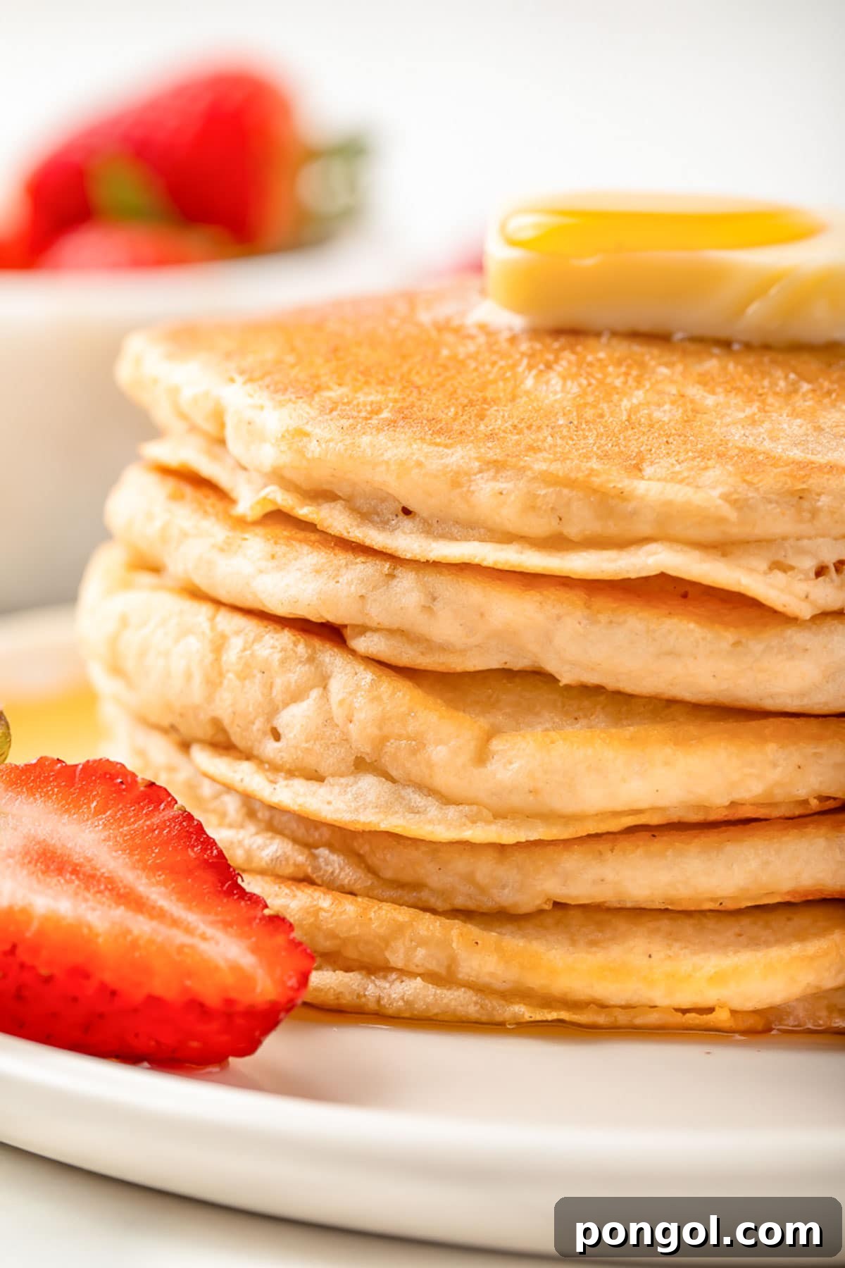 A stack of fluffy, gluten-free pancakes on a white plate with a halved strawberry and a drizzle of syrup.