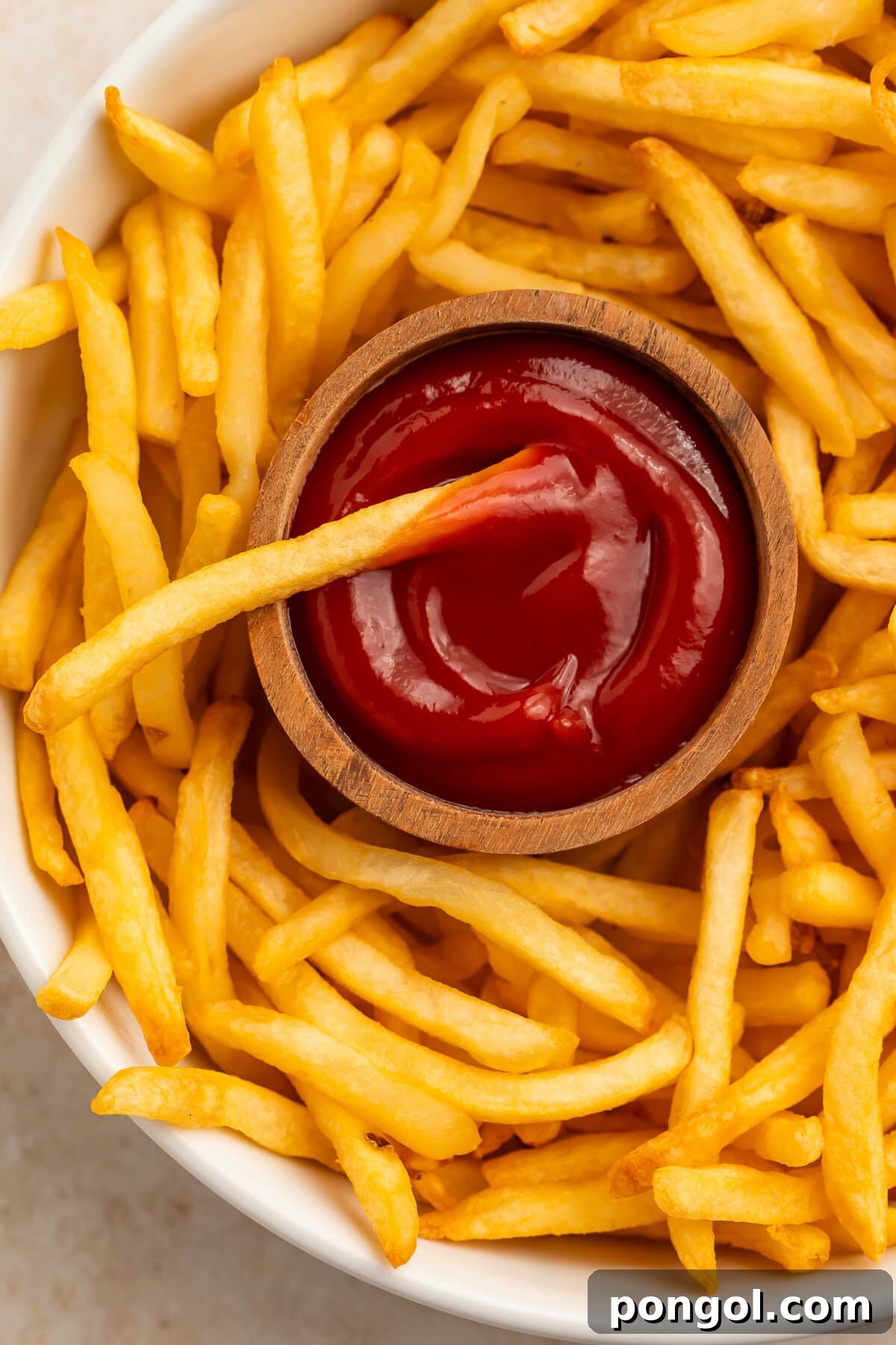 Golden air fryer frozen french fries on a white plate with ketchup in a wooden bowl.