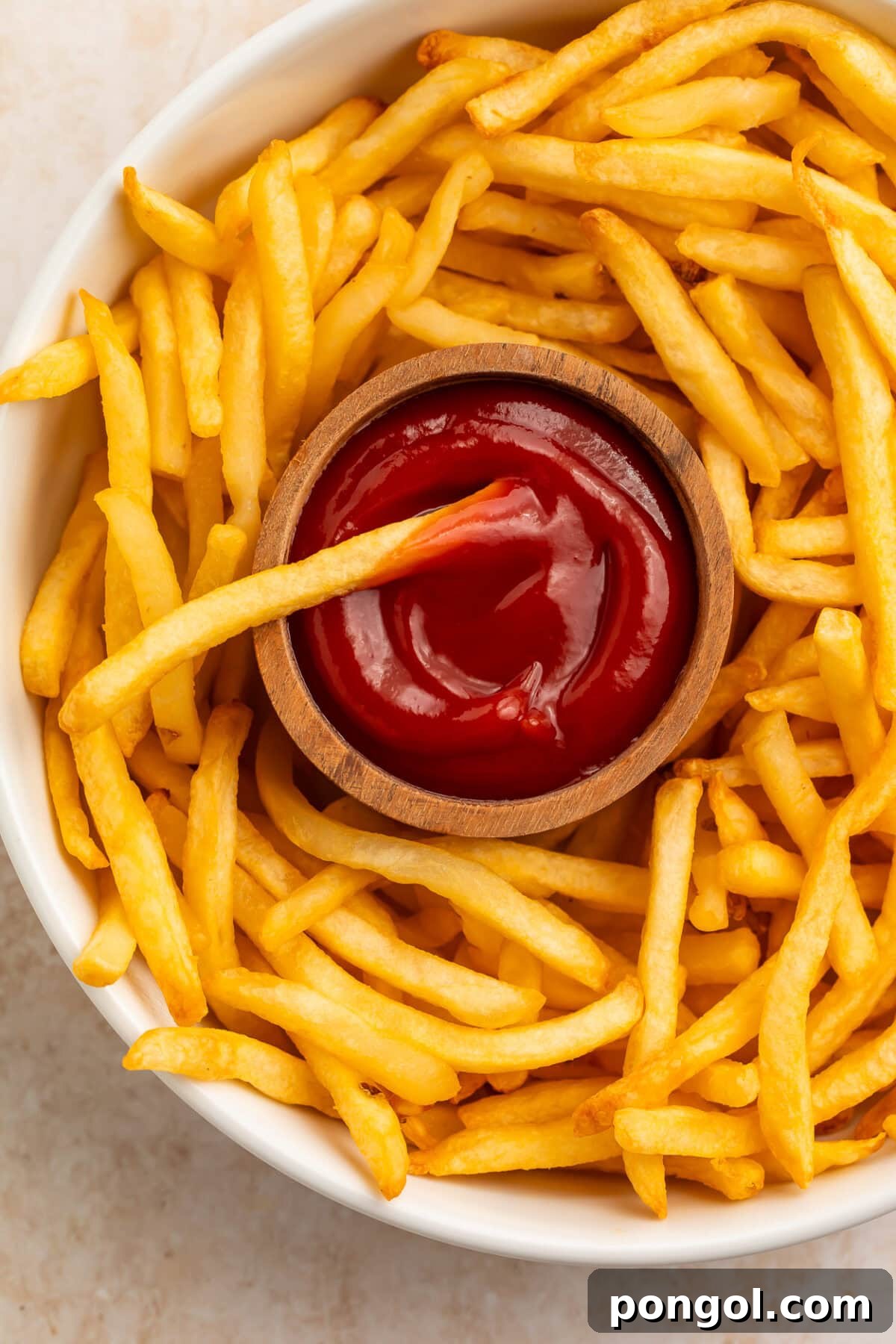 Crispy golden french fries surrounding a wooden bowl of ketchup on a white plate.