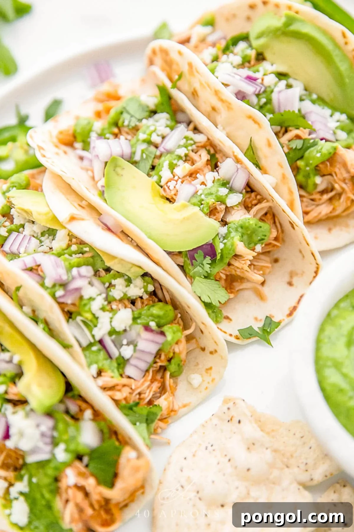 Crockpot chicken tacos arranged on a white plate. The tacos are topped with diced red onion, slices of avocado, and crumbled cheese.