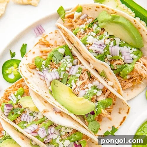 Crockpot chicken tacos arranged on a white plate. The tacos are topped with diced red onion, slices of avocado, and crumbled cheese.