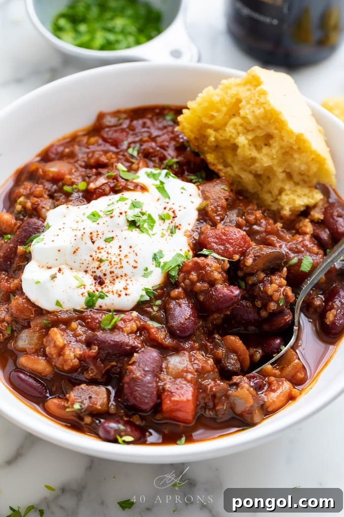 A hearty bowl of rich, dark vegan chili, garnished with fresh cilantro and a swirl of vegan sour cream, looking utterly delicious and satisfying.