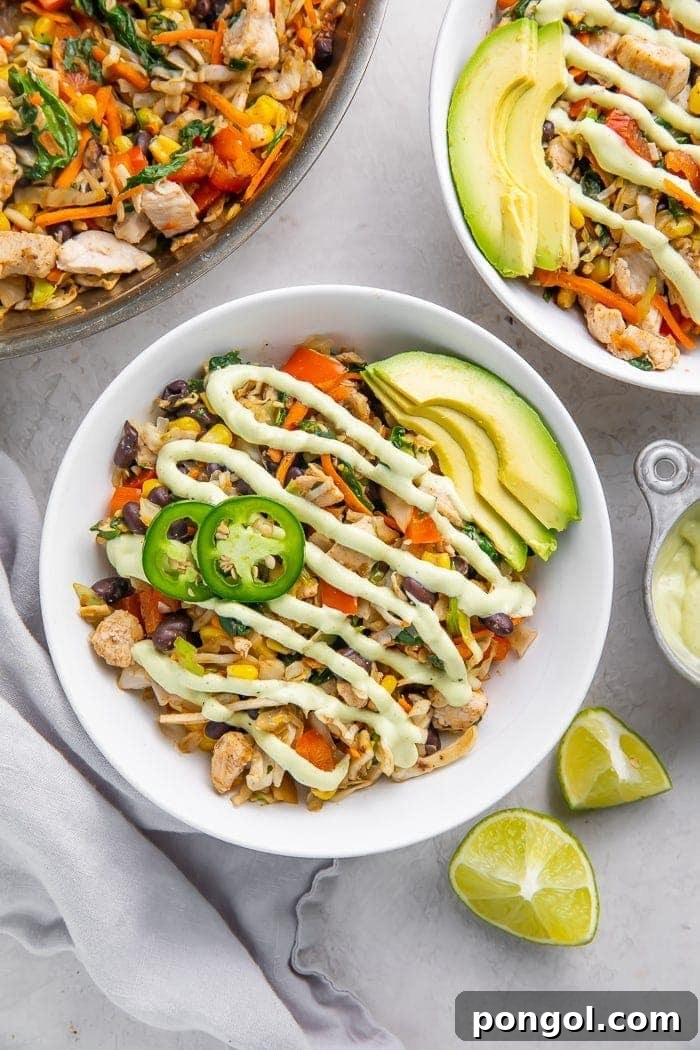 A vibrant bowl of Southwestern egg roll ingredients, including chicken, black beans, corn, and bell peppers, drizzled with creamy avocado ranch dressing, garnished with fresh lime wedges on a rustic surface.