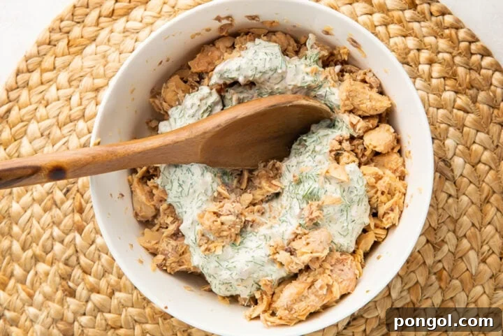 Creamy dill sauce added to a bowl of canned salmon salad with a wooden spoon.